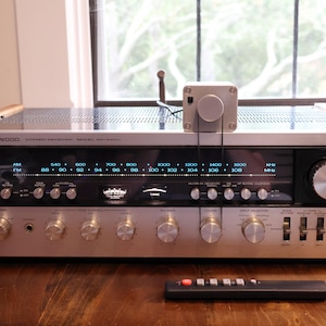 K&ouml;nnte beinhalten: Ein Vintage-Stereo-Receiver von Kenwood mit silberner Front, Holzseitenteilen und zahlreichen Kn&ouml;pfen. Obenauf befindet sich ein kleiner silberner Kasten mit einem Drehknopf, der mit Dr&auml;hten verbunden ist. Eine schwarze Fernbedienung liegt davor.