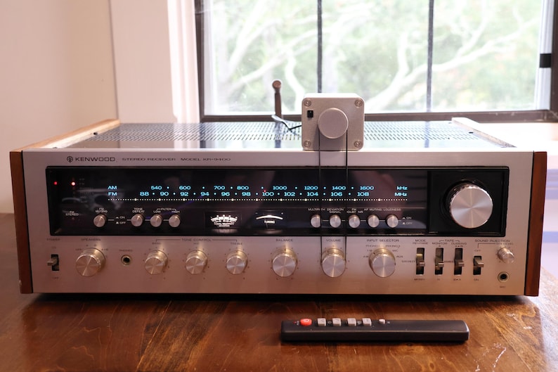 K&ouml;nnte beinhalten: Ein Vintage-Stereo-Receiver von Kenwood mit silberner Front, Holzseitenteilen und zahlreichen Kn&ouml;pfen. Obenauf befindet sich ein kleiner silberner Kasten mit einem Drehknopf, der mit Dr&auml;hten verbunden ist. Eine schwarze Fernbedienung liegt davor.