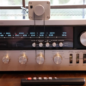 K&ouml;nnte beinhalten: Silberner Vintage-Stereo-Receiver mit mehreren Kn&ouml;pfen und Tasten sowie einer Digitalanzeige der Radiofrequenzen. Ein kleines silbernes Ger&auml;t ist oben montiert. Eine schwarze Fernbedienung befindet sich im Vordergrund. Modell KR-9400.