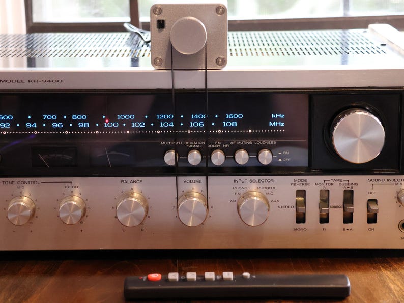 K&ouml;nnte beinhalten: Silberner Vintage-Stereo-Receiver mit mehreren Kn&ouml;pfen und Tasten sowie einer Digitalanzeige der Radiofrequenzen. Ein kleines silbernes Ger&auml;t ist oben montiert. Eine schwarze Fernbedienung befindet sich im Vordergrund. Modell KR-9400.
