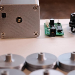 K&ouml;nnte beinhalten: Ein silberfarbenes rechteckiges Ger&auml;t mit einem Netzschalter und einem zentralen Anschluss, begleitet von einer gr&uuml;nen Leiterplatte, einem schwarzen Netzkabel und mehreren silbernen kreisf&ouml;rmigen Komponenten mit zentralen Pfosten. Die Artikel sind auf einer hellen Oberfl&auml;che angeordnet.