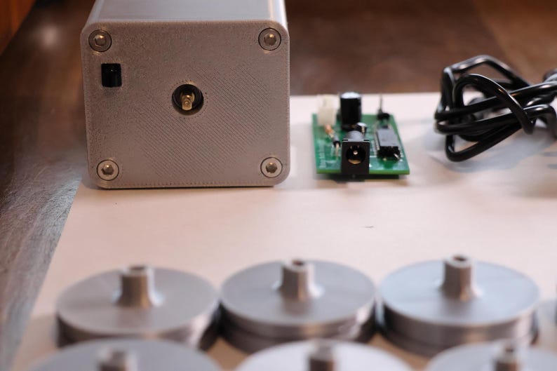 K&ouml;nnte beinhalten: Ein silberfarbenes rechteckiges Ger&auml;t mit einem Netzschalter und einem zentralen Anschluss, begleitet von einer gr&uuml;nen Leiterplatte, einem schwarzen Netzkabel und mehreren silbernen kreisf&ouml;rmigen Komponenten mit zentralen Pfosten. Die Artikel sind auf einer hellen Oberfl&auml;che angeordnet.