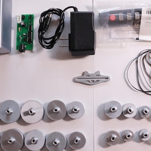 K&ouml;nnte beinhalten: Eine Ansammlung elektronischer Komponenten, darunter eine silberne Box, eine gr&uuml;ne Leiterplatte, ein schwarzes Netzteil und mehrere silberne Spulen. Ebenfalls enthalten sind eine Fernbedienung, ein graues Kunststoffteil und ein schwarzes Kabel.