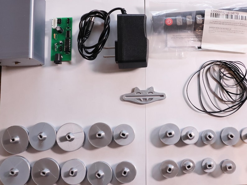 K&ouml;nnte beinhalten: Eine Ansammlung elektronischer Komponenten, darunter eine silberne Box, eine gr&uuml;ne Leiterplatte, ein schwarzes Netzteil und mehrere silberne Spulen. Ebenfalls enthalten sind eine Fernbedienung, ein graues Kunststoffteil und ein schwarzes Kabel.
