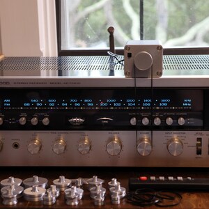 K&ouml;nnte beinhalten: Ein Vintage-Stereo-Receiver von Kenwood mit silberner Front, zahlreichen Kn&ouml;pfen und einer Digitalanzeige. Der Receiver hat eine Holzoberseite und wird von einer Fernbedienung und mehreren silberfarbenen Komponenten begleitet. Oben ist eine kleine silberne Box befestigt.