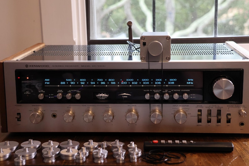 K&ouml;nnte beinhalten: Ein Vintage-Stereo-Receiver von Kenwood mit silberner Front, zahlreichen Kn&ouml;pfen und einer Digitalanzeige. Der Receiver hat eine Holzoberseite und wird von einer Fernbedienung und mehreren silberfarbenen Komponenten begleitet. Oben ist eine kleine silberne Box befestigt.
