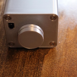 K&ouml;nnte beinhalten: Nahaufnahme eines silberfarbenen elektronischen Ger&auml;ts mit einem gro&szlig;en, runden Knopf und einem kleinen Netzschalter. Das rechteckige Ger&auml;t hat Schrauben an jeder Ecke. Der Hintergrund ist eine Holzoberfl&auml;che.