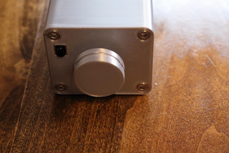K&ouml;nnte beinhalten: Nahaufnahme eines silberfarbenen elektronischen Ger&auml;ts mit einem gro&szlig;en, runden Knopf und einem kleinen Netzschalter. Das rechteckige Ger&auml;t hat Schrauben an jeder Ecke. Der Hintergrund ist eine Holzoberfl&auml;che.