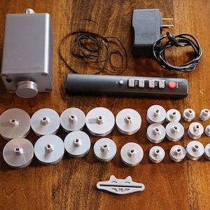K&ouml;nnte beinhalten: Eine Ansammlung silberfarbener elektronischer Komponenten, darunter ein rechteckiger Kasten mit einem Knopf, eine Fernbedienung und mehrere kreisf&ouml;rmige Teile. Ein Netzteil und ein kleines wei&szlig;es Kunststoffteil sind ebenfalls vorhanden.
