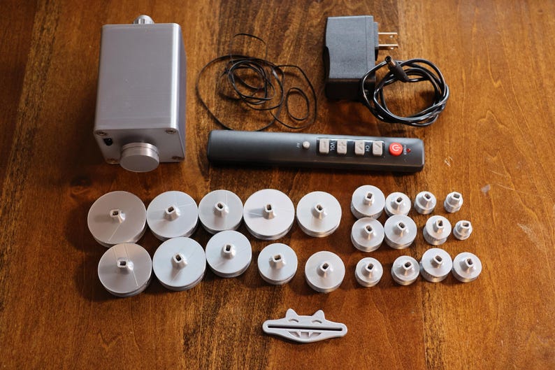 K&ouml;nnte beinhalten: Eine Ansammlung silberfarbener elektronischer Komponenten, darunter ein rechteckiger Kasten mit einem Knopf, eine Fernbedienung und mehrere kreisf&ouml;rmige Teile. Ein Netzteil und ein kleines wei&szlig;es Kunststoffteil sind ebenfalls vorhanden.