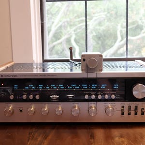 K&ouml;nnte beinhalten: Ein Vintage-Stereo-Receiver von Kenwood mit silberner Front, Holzseitenteilen und zahlreichen Kn&ouml;pfen. Der Receiver zeigt Radiofrequenzen an und hat einen kleinen, silbernen Steuerkasten oben. Das Ger&auml;t steht auf einer Holzoberfl&auml;che.