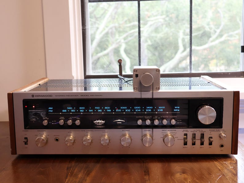 K&ouml;nnte beinhalten: Ein Vintage-Stereo-Receiver von Kenwood mit silberner Front, Holzseitenteilen und zahlreichen Kn&ouml;pfen. Der Receiver zeigt Radiofrequenzen an und hat einen kleinen, silbernen Steuerkasten oben. Das Ger&auml;t steht auf einer Holzoberfl&auml;che.