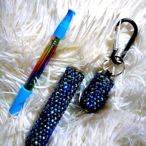 May include: Two small, blue, jeweled containers. One is attached to a black carabiner with a silver ring. The other is next to a blue and rainbow-colored pen with a blue tip.