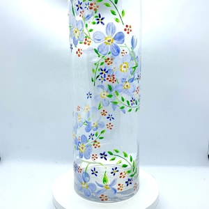May include: A clear glass vase with hand-painted floral designs. The vase features light blue flowers, green vines, and small orange and red accents. The cylindrical vase is approximately 20 cm tall.