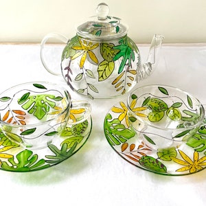 May include: A hand-painted glass teapot and two teacups with matching saucers. The teapot and cups feature a green and yellow floral design with leaves and flowers.