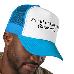 May include: A blue and white trucker hat with the text "Friend of Dorothy (Zbornak)". The hat has a mesh back and a curved brim. The hat is worn by a person wearing a tan button-down shirt.