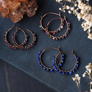 May include: Three pairs of copper hoop earrings with gemstone beads. The earrings are on a dark blue surface. The earrings are decorated with red, pink, and blue gemstones.