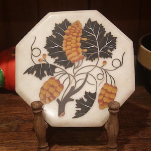 May include: An octagonal trivet with a grape vine design. The trivet is white with a black and orange grape vine illustration. It is displayed on a wooden stand. A black bowl with a tan rim is in the background.