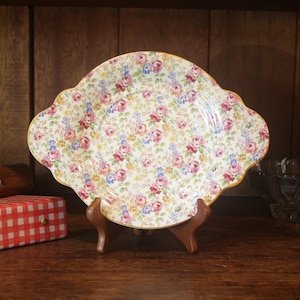 May include: A white ceramic plate with a floral pattern in shades of pink, yellow, and blue. The plate has a scalloped edge and is displayed on a wooden stand.