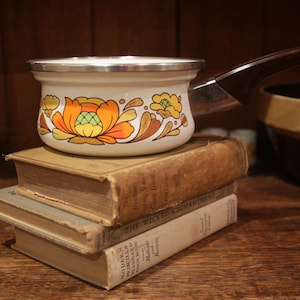 Pode incluir: Uma panela de esmalte branco com um padrão floral vibrante em tons de laranja e amarelo no exterior, uma borda de metal prateado e uma alça castanho-escura, pousada sobre uma pilha de dois livros de capa dura desgastados.
