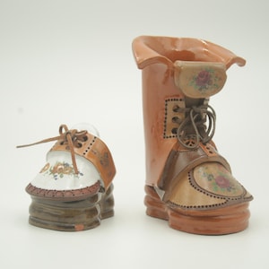 May include: Two ceramic boot-shaped vases with floral designs. The larger boot is brown with a light brown leather-like texture and a brown lace. The smaller boot is brown with a white top and a brown lace.