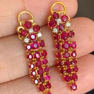 May include: A pair of gold drop earrings featuring a floral top design set with vibrant red round-cut stones and small clear accent stones. The elongated body of each earring consists of multiple rows of faceted red stones in a tapered, vertical arrangement.