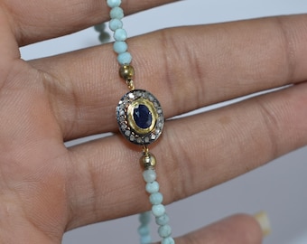 Può includere: Un delicato braccialetto con piccole perline azzurre infilate insieme. Il braccialetto ha un pendente centrale ovale in oro con una gemma blu scuro circondata da pietre più piccole e trasparenti. Il braccialetto è tenuto su uno sfondo neutro.