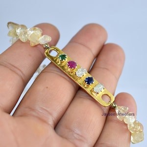 Può includere: Un braccialetto color oro con una barra rettangolare con cinque pietre preziose colorate: verde, rosso, bianco e blu. Il braccialetto è collegato a un filo di perle gialle chiare di forma irregolare. Il braccialetto è tenuto in una mano.