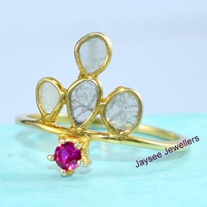 Puede incluir: Un anillo dorado con cuatro piedras claras de forma irregular y una pequeña gema roja redonda. El anillo está sobre una superficie azul claro, con el texto "Jaysee Jewellers" visible.