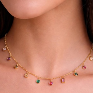 Op de afbeelding: Een delicate gouden ketting met een reeks kleurrijke edelstenen. De ketting heeft een fijne ketting met kleine, bungelende edelstenen in verschillende vormen en kleuren, waaronder roze, groene en heldere stenen. De ketting wordt om de nek gedragen.