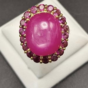 May include: A gold ring with a large, oval magenta gemstone, encircled by smaller pink gemstones. The ring is displayed on a white surface against a dark backdrop. The gemstones are the main feature of the jewellery.