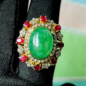 May include: A gold ring with a large oval green gemstone, encircled by smaller red gemstones and a halo of clear stones. The ring is set against a black background.