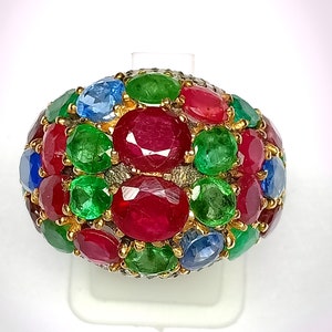 May include: A gold ring set with a cluster of oval-shaped gemstones. The ring features a mix of red rubies, green emeralds, and blue sapphires.