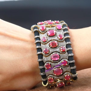 May include: A wide silver bangle bracelet with a black fabric band. The bracelet is adorned with a row of oval-shaped pink gemstones and smaller clear gemstones.