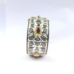 Könnte beinhalten: Ein silbernes Armreif-Armband mit geometrischem Design und einem roten Edelstein, der in einen goldenen Rahmen gefasst ist. Das Armband ist mit kleinen Diamanten verziert.