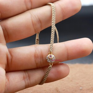 May include: A beige string bracelet with a gold charm featuring a pink gemstone surrounded by clear rhinestones.