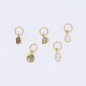 May include: Five gold pendants, each featuring a raw-cut gemstone in shades of brown, grey, and clear. The gemstones are set in gold bezels with a small loop. The pendants are displayed against a white background. The gemstones are in various shapes.