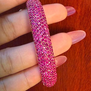May include: A dazzling pink bracelet, encrusted with numerous small, glittering gemstones. The bracelet is a vivid fuchsia hue, composed of many tiny, round stones. The jewellery is held in a hand with long, polished nails.