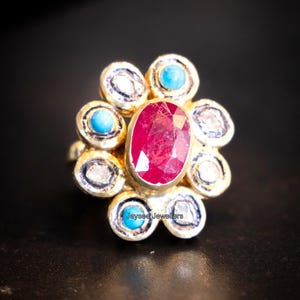 May include: A gold ring with a central oval ruby, surrounded by a floral arrangement of diamonds and turquoise stones. The ring has the text "Jayseed Jewellers". The ring is set against a black background.