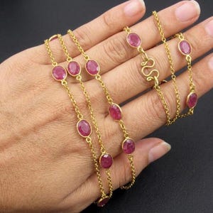 Op de afbeelding: Een goudkleurige ketting met ovale, dieprode edelstenen. De edelstenen zijn gelijkmatig verdeeld over de fijne ketting. De ketting wordt op een hand getoond, waarbij het ontwerp en de kleur worden benadrukt.