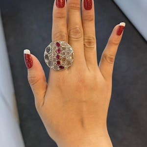 Può includere: Un grande anello rotondo d'argento con un gruppo di pietre preziose. L'anello presenta un mix di pietre chiare e rosse disposte in un motivo circolare. L'anello è mostrato su una mano con smalto rosso.