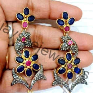 Puede incluir: Un par de pendientes de oro con piedras preciosas azules y rosas. Los pendientes tienen forma de flores con un delicado diseño floral. Los pendientes están engastados con pequeños diamantes.