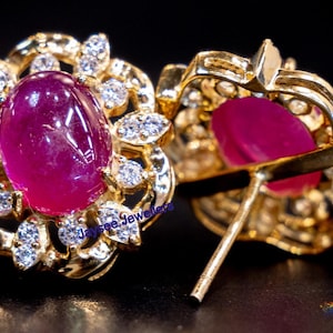 以下が含まれることがあります： 大きな楕円形のルビーが、小さな白い宝石で飾られた花柄にセットされた、ゴールドのイヤリングのペア。イヤリングはポストとバタフライバックです。