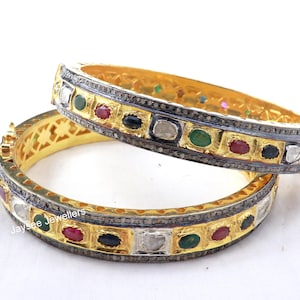 Könnte beinhalten: Zwei goldene Armreifen mit silbernem und schwarzem Besatz. Die Armreifen sind mit einer Reihe von bunten Edelsteinen verziert, darunter rot, grün, blau und weiß.