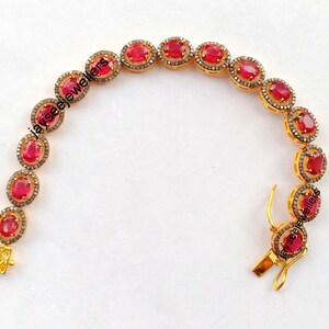 May include: A gold bracelet with a row of oval-shaped red gemstones surrounded by smaller clear gemstones. The bracelet has a clasp closure.