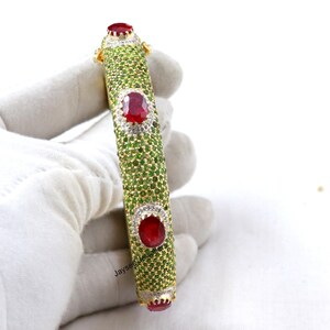 Puede incluir: Un brazalete de oro con piedras preciosas rojas y verdes. El brazalete está engastado con piedras redondas rojas y piedras verdes más pequeñas. El brazalete está sobre una superficie blanca.