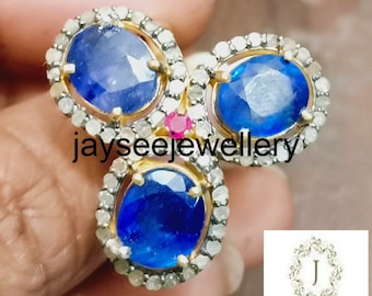 May include: A gold ring featuring three oval-shaped blue sapphires, each encircled by a halo of small diamonds. A small red ruby is set between the sapphires.