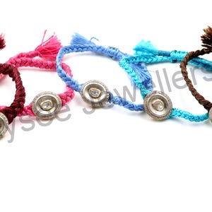May include: A collection of braided bracelets in various colors: burgundy, pink, light blue, turquoise, and brown. Each bracelet features a silver-toned circular centerpiece with a clear stone and a matching tassel.
