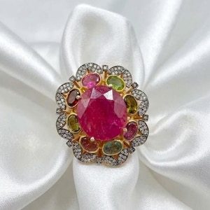 Puede incluir: Un anillo de oro con una gran piedra central redonda y facetada de color rosa, rodeada de gemas ovaladas multicolores y un borde festoneado de pequeñas piedras blancas engastadas en pavé, creando un diseño floral de varias capas.