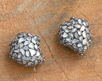 May include: Two silver-toned, irregular-shaped earrings. Each earring is encrusted with numerous small, irregularly shaped, clear stones. The earrings are displayed on a textured, brown wooden surface.